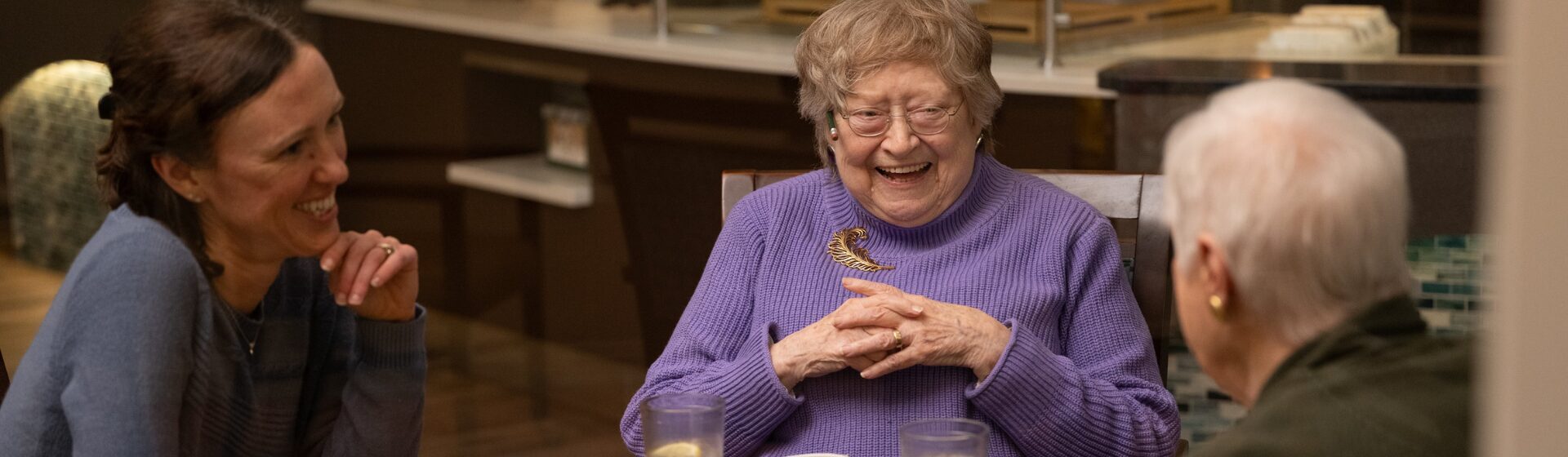 Three residents laughing and sharing a meal together at a table in the community dining room, highlighting the enjoyable food service on-site amenities.