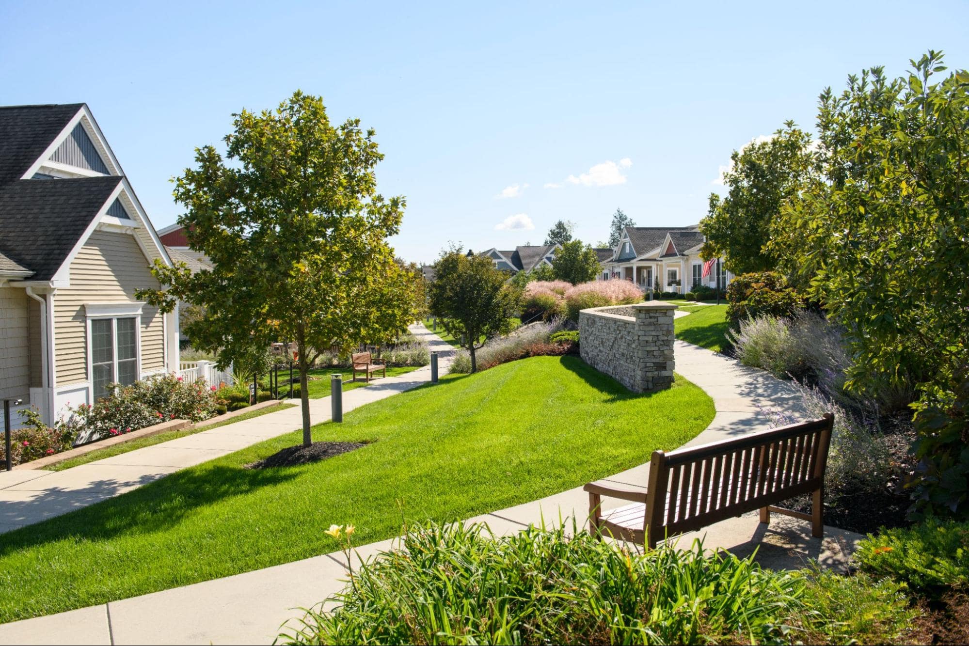 A wooden bench sits next to a winding sidewalk and green landscaping in a peaceful independent living neighborhood.