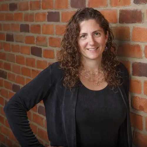 A portrait of a smiling woman with curly brown hair wearing a black outfit, standing in front of a red brick wall.