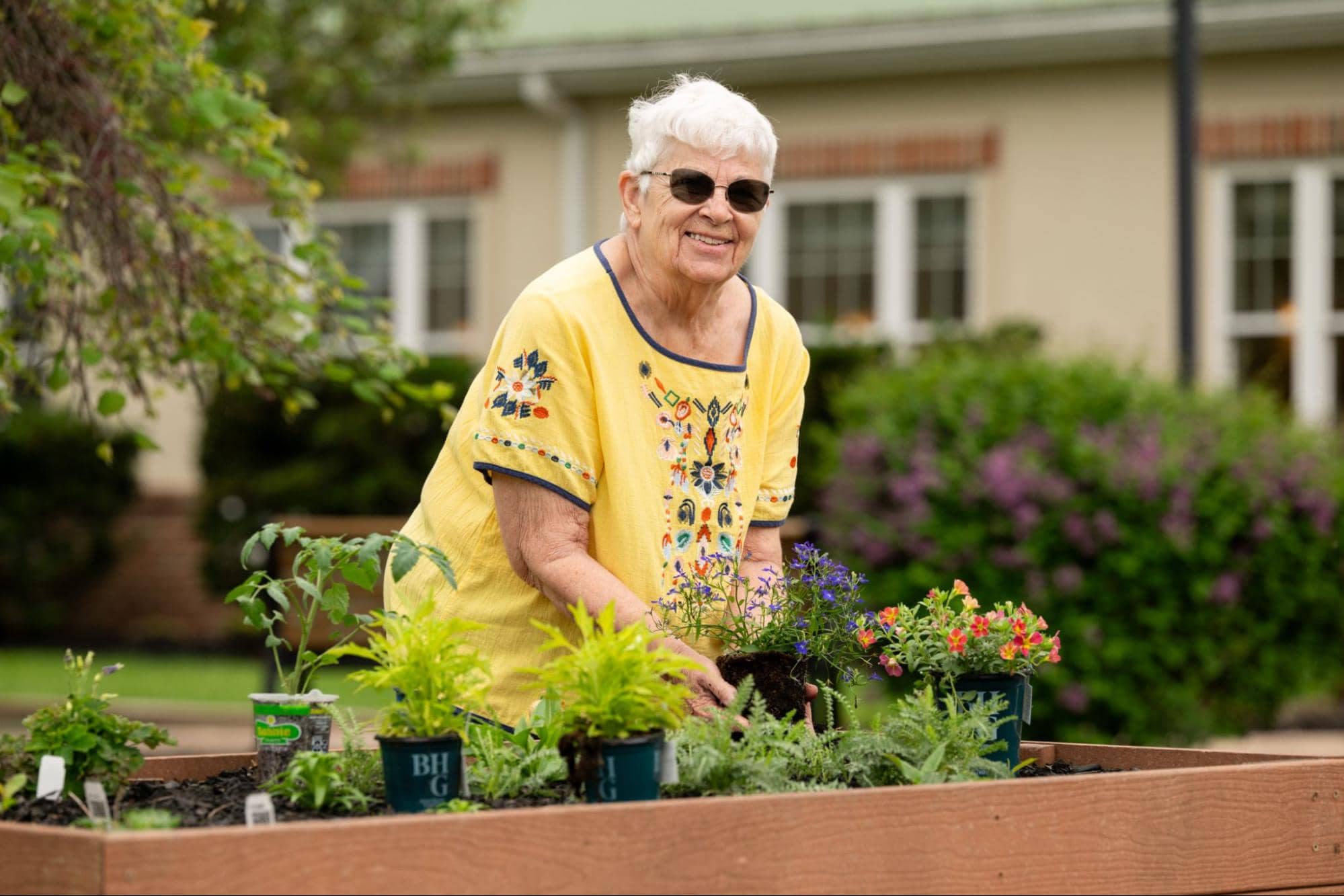 A smiling older woman with sunglasses and a yellow shirt plants flowers in a raised garden bed, enjoying the low-maintenance outdoor amenities that come with rightsizing in retirement.