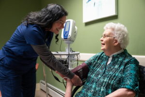 A friendly caregiver smiles while checking an older woman's blood pressure on a comfortable couch, highlighting the dedicated support available when exploring Personal Care vs. Assisted Living.