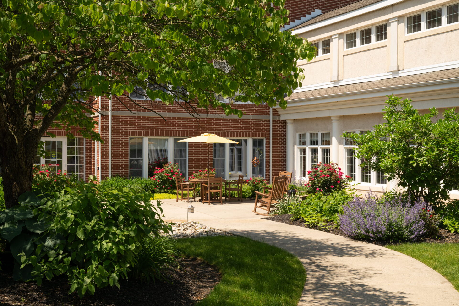 A sunny courtyard at Frederick Living showcasing premier campus amenities, including comfortable wooden rocking chairs, a patio dining set with a yellow umbrella, and manicured floral gardens.