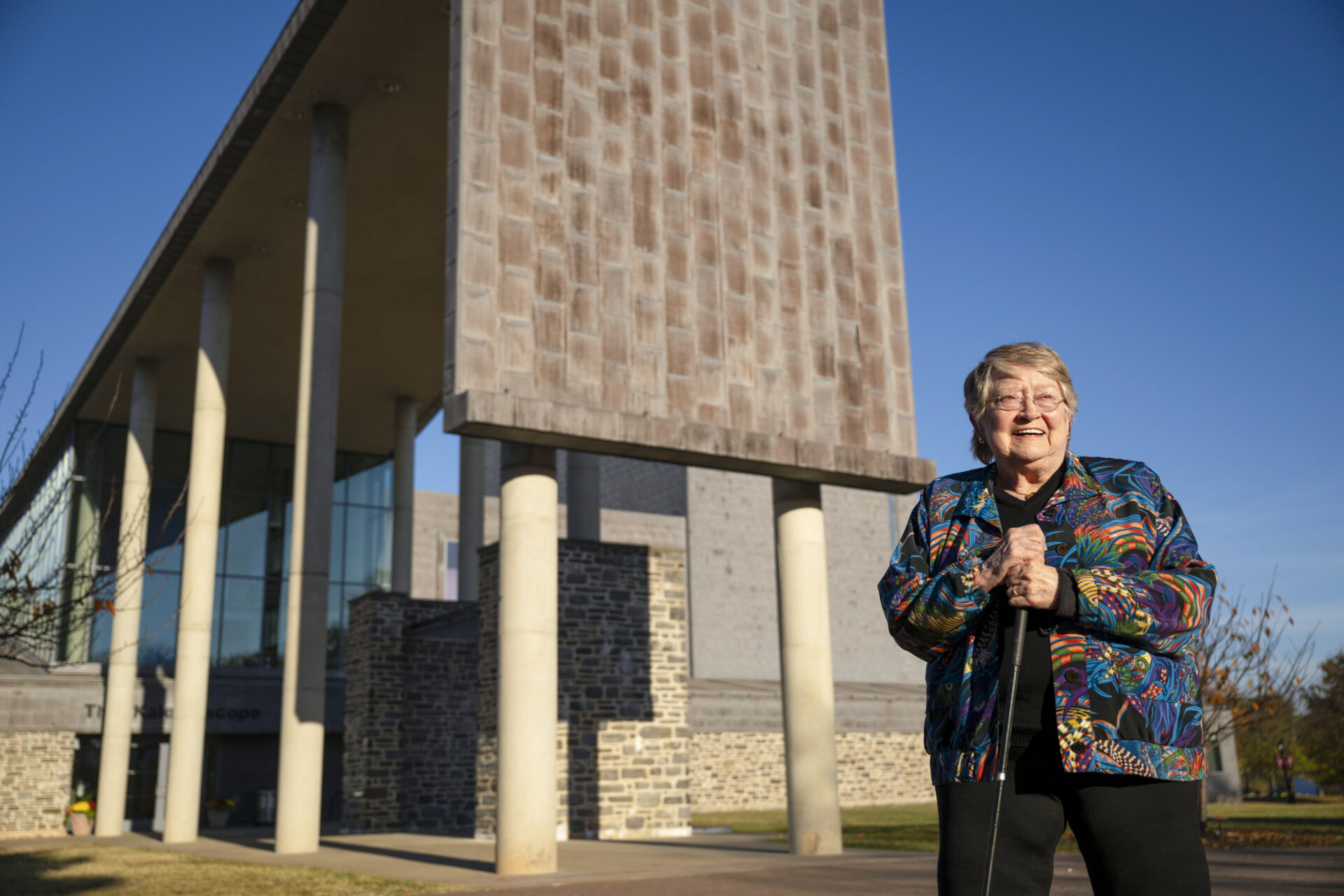 A smiling woman in a colorful jacket standing outside a modern campus building, illustrating the lifestyle and peace of mind secured by a retirement community entrance fee.