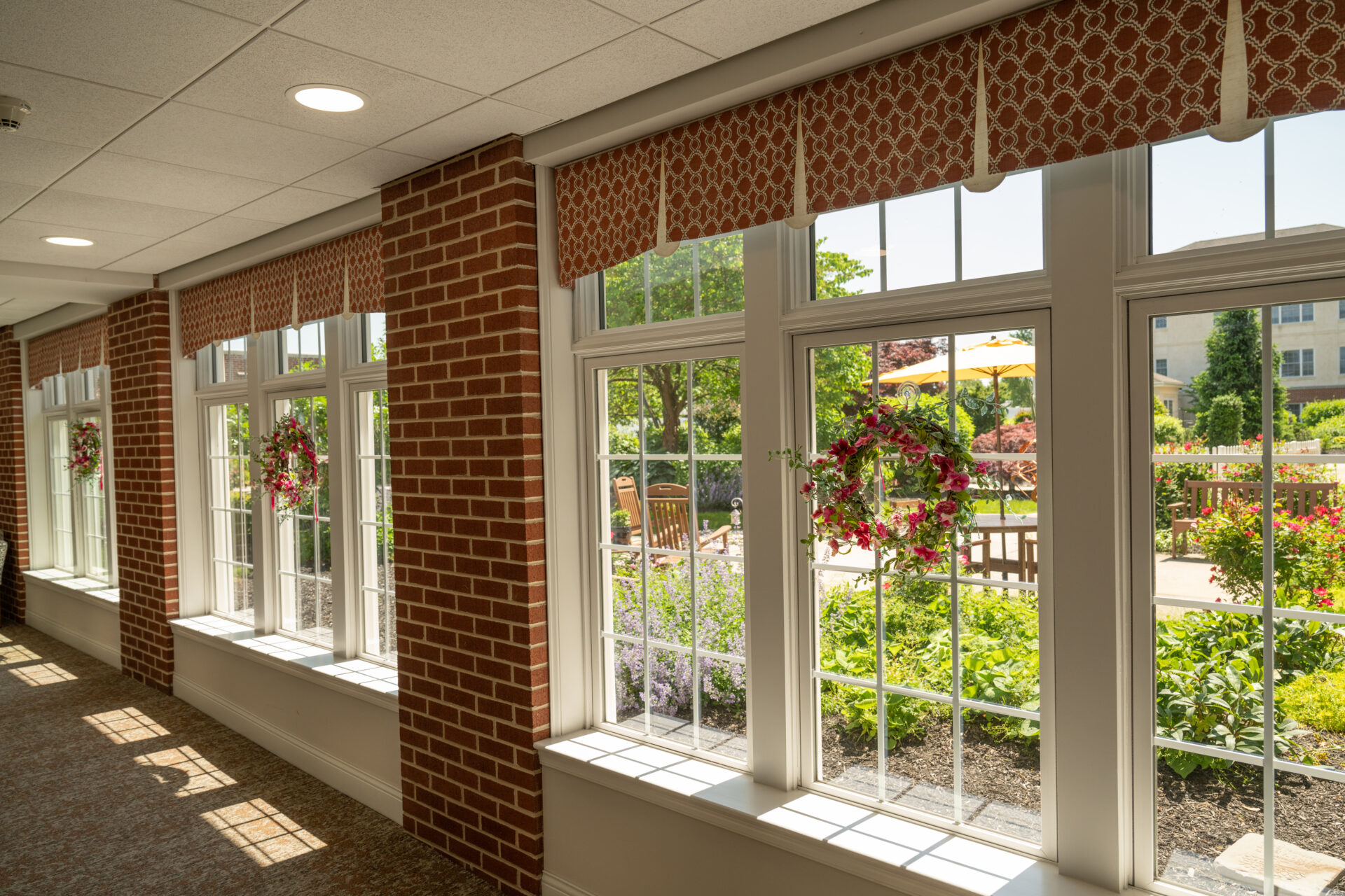 A sunlit hallway with brick accents and large windows overlooking a lush garden courtyard, illustrating the high-quality environment included in retirement community pricing.