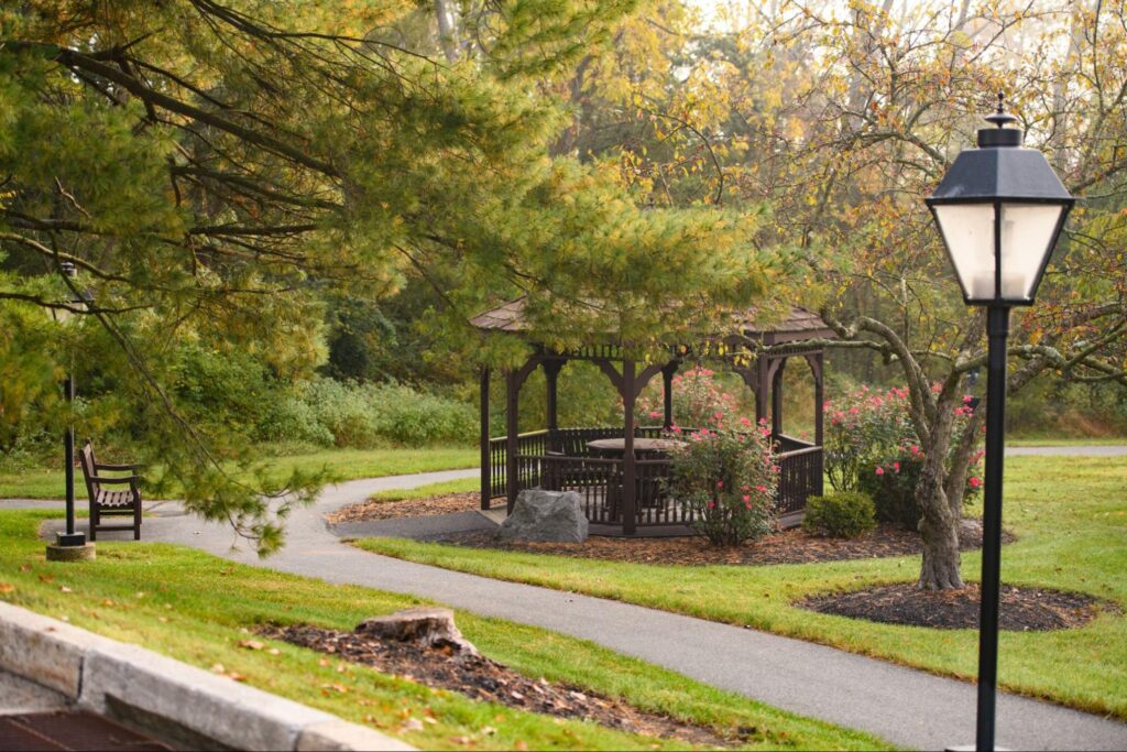 A scenic and peaceful outdoor park area at a facility offering long term care options, featuring a winding paved walking path, a wooden gazebo with patio seating, a park bench, and a classic black lamppost surrounded by mature trees and lush green grass.
