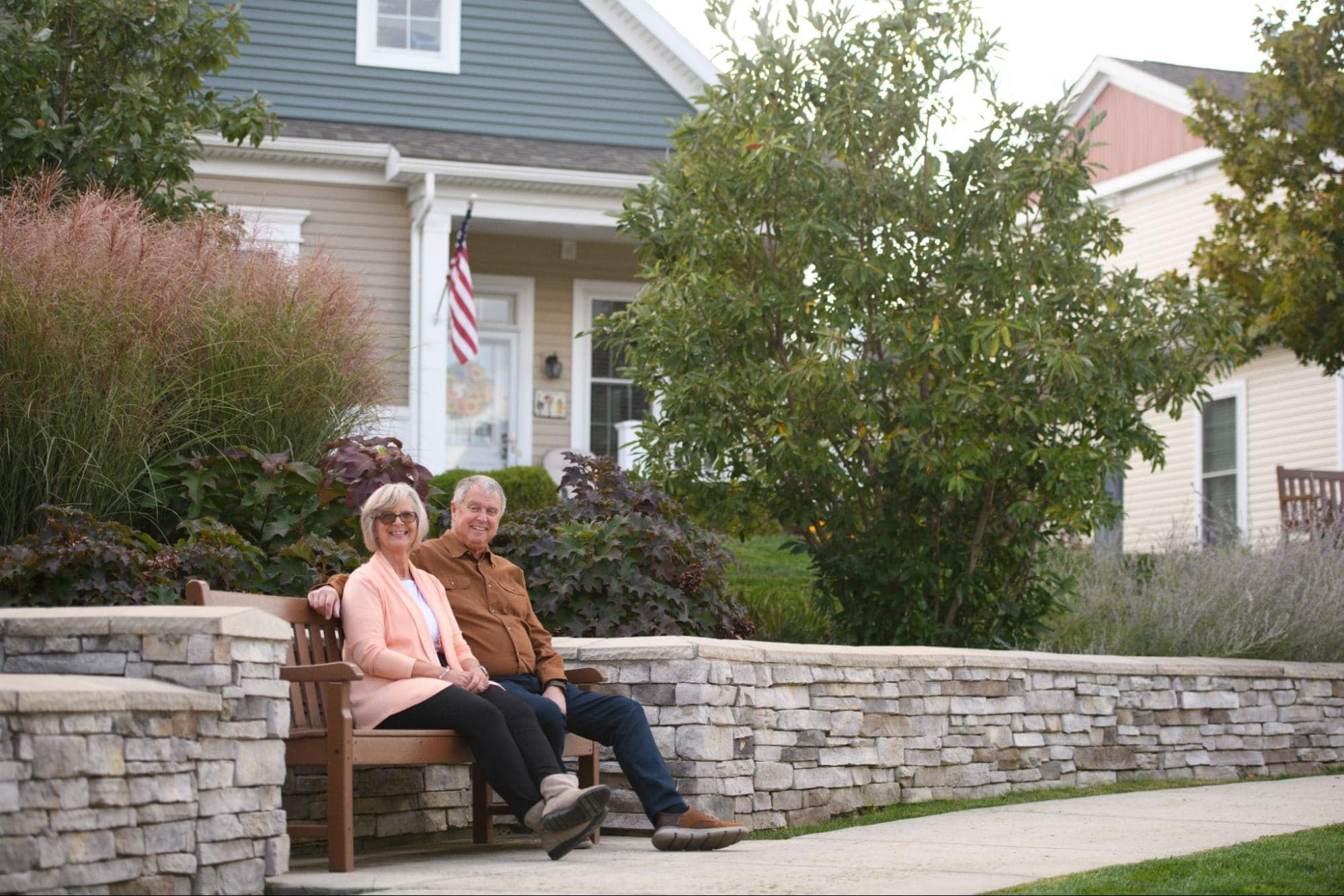 A senior man and woman smile while sitting together on a bench outside beautiful cottage homes in an active retirement community.