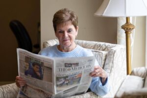 An older woman relaxes in a comfortable armchair by a lamp, experiencing stress-free living while reading "The Mercury" newspaper.
