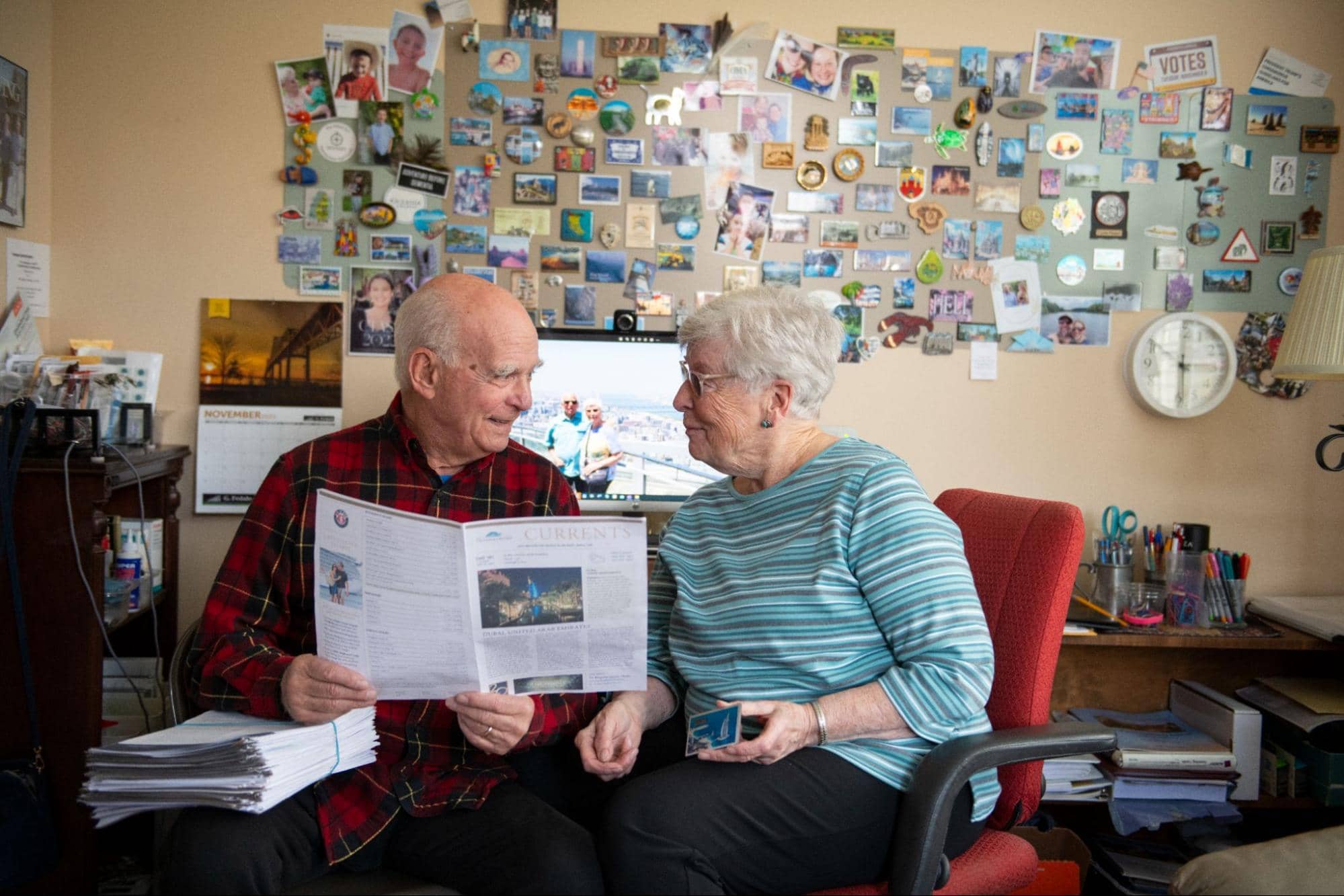 A senior couple smiles at each other in their community apartment while holding a newsletter, sitting in front of a wall covered in cherished family photos during their transition to senior living.
