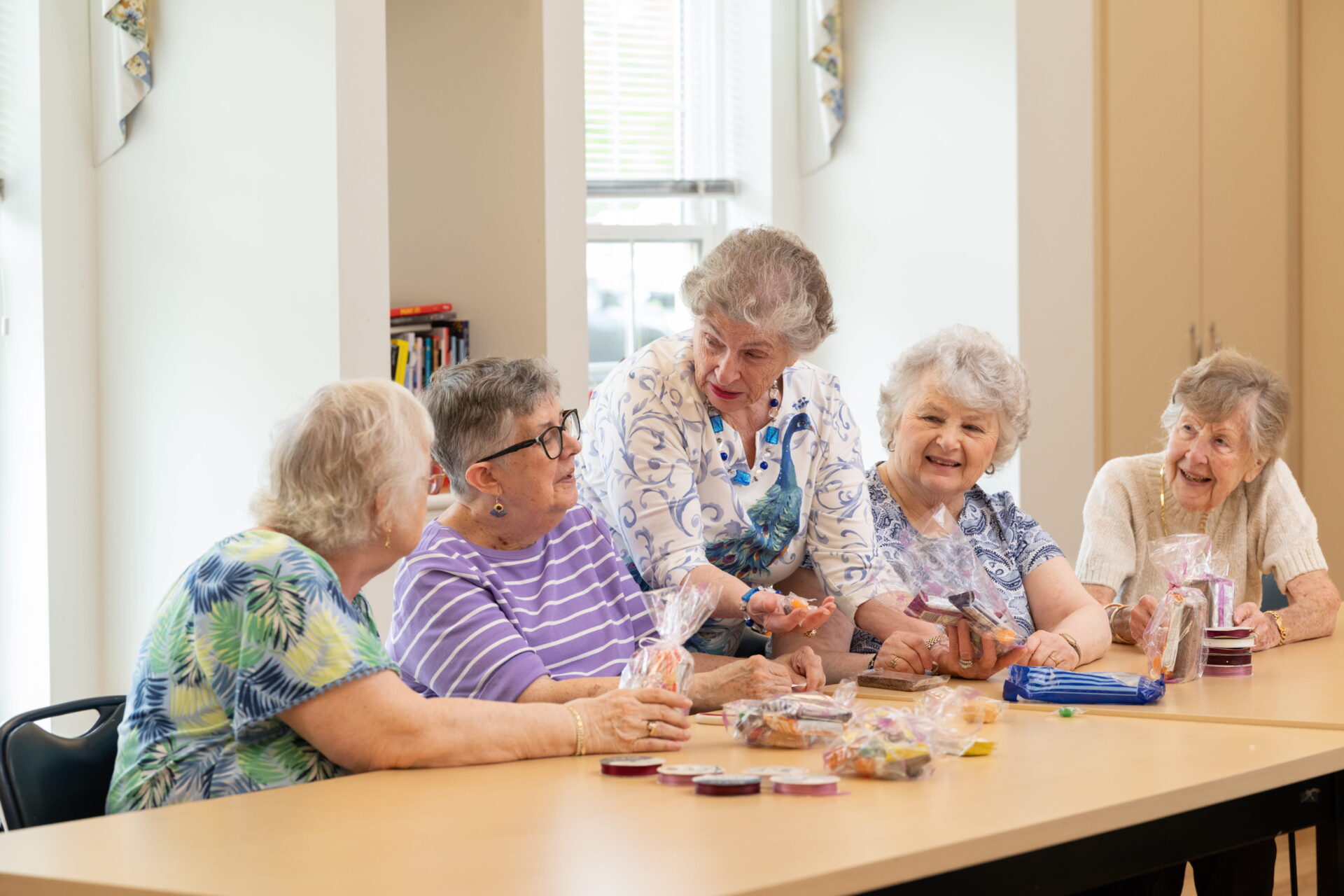 A group of five senior women happily chatting and interacting while sitting around a long wooden table during a social activity in a bright common room at a continuing care retirement community.