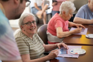 A smiling woman sits at a table participating in arts and crafts as part of her memory care program, supported by a staff member assisting with activities of daily care.