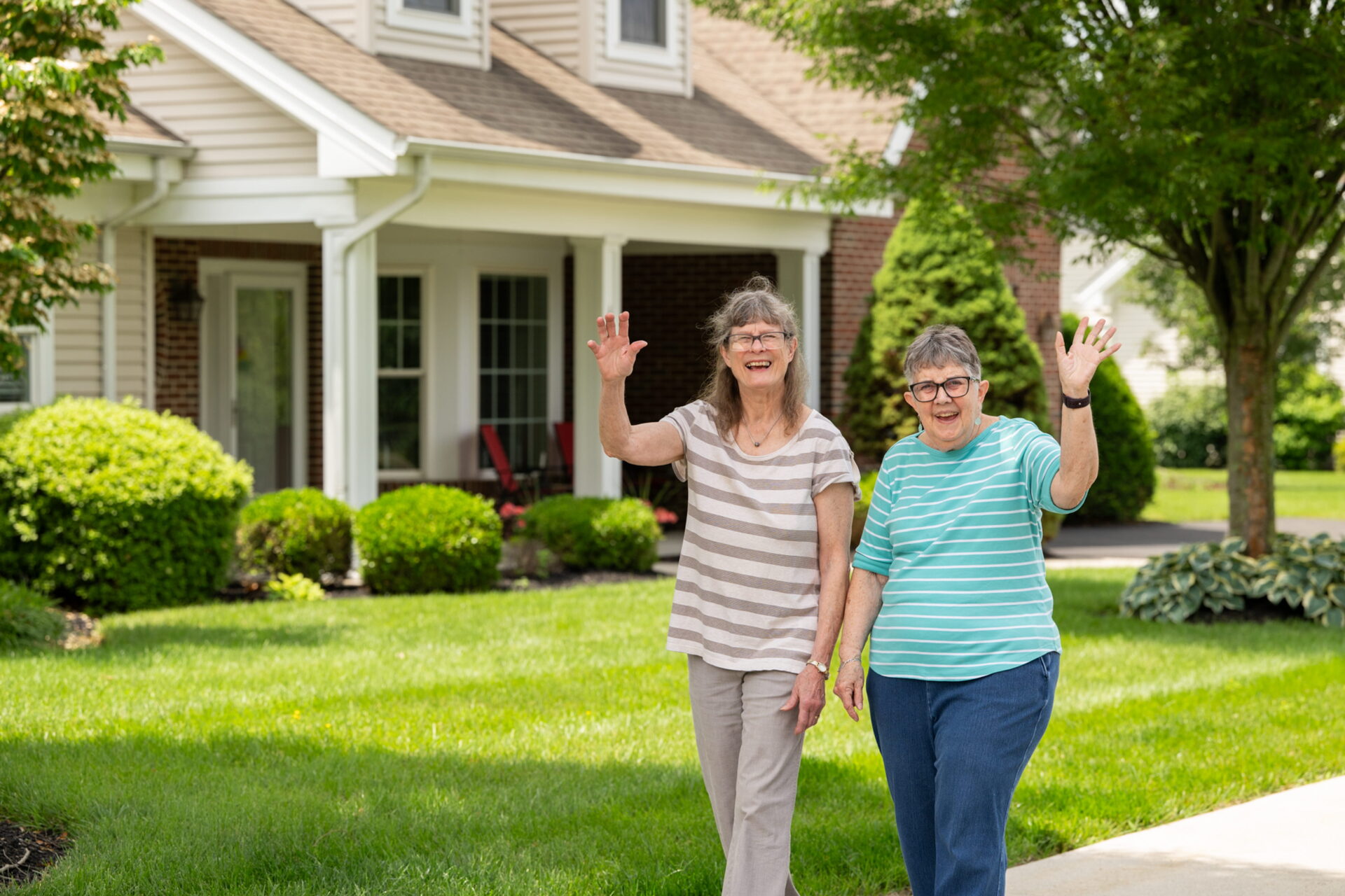 Two cheerful senior women, both waving friendly hellos and wearing striped shirts, walk together on a sidewalk in front of a welcoming, well-maintained house. The scene represents active retirement living in a "55 and over communities in collegeville pa" environment, featuring a lush green lawn and manicured landscaping under a clear sky.