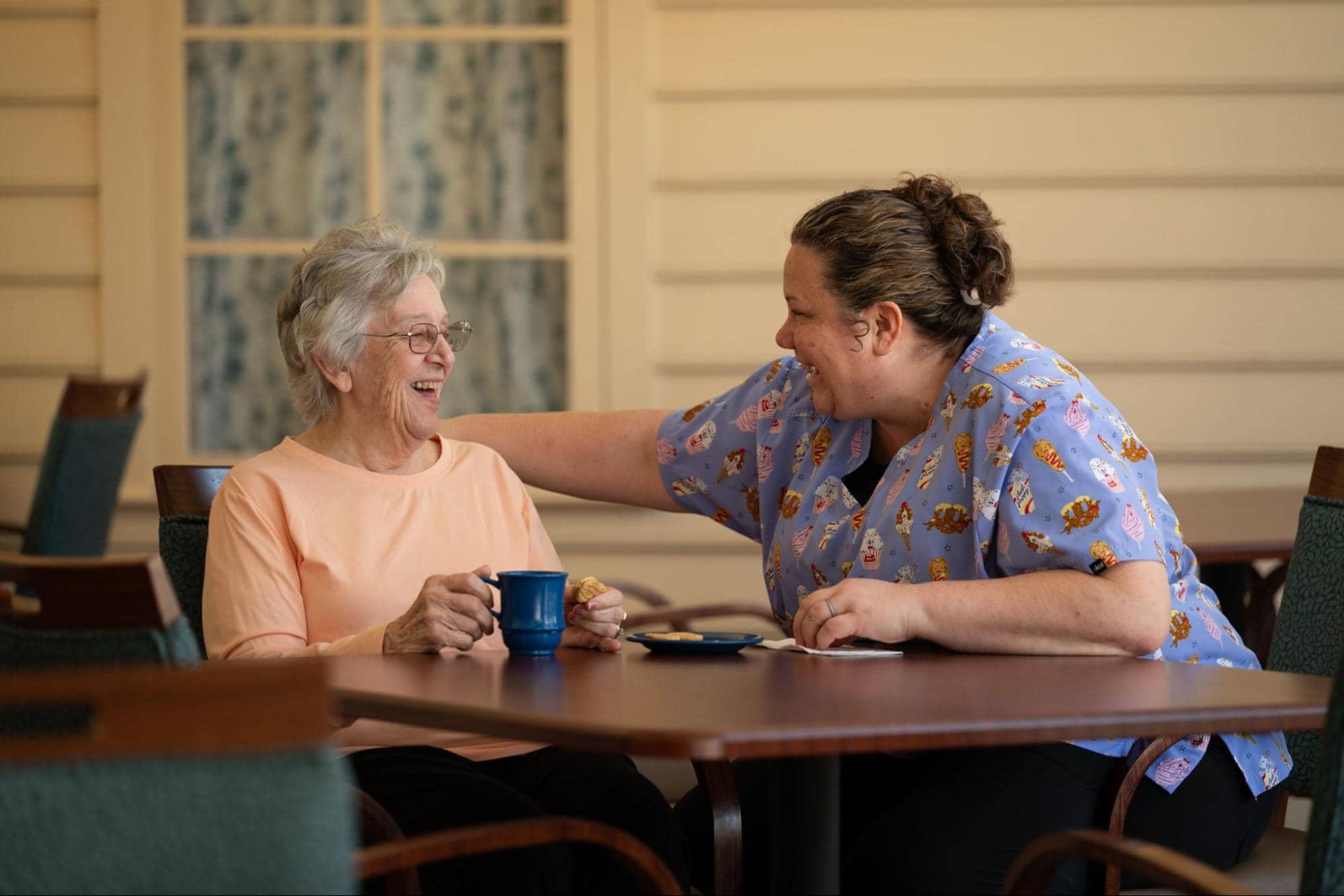 An interior photo showing an elderly resident in a peach shirt laughing joyfully as she sits at a wooden table, holding a blue coffee cup, with a smiling caregiver in a blue patterned uniform placing a hand on her shoulder, illustrating quality companionship care for your loved one.